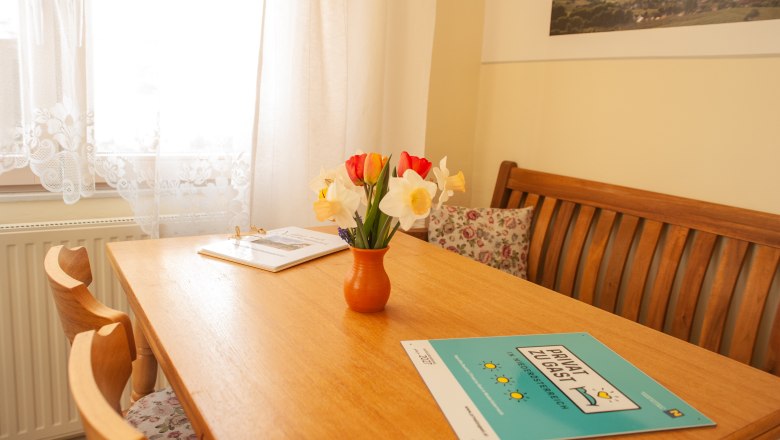 Dining area, © Fam. Heinrich A cozy dining area with a wooden table, a bouquet of flowers in a vase and brochures. Light-colored curtains and a picture on the wall.