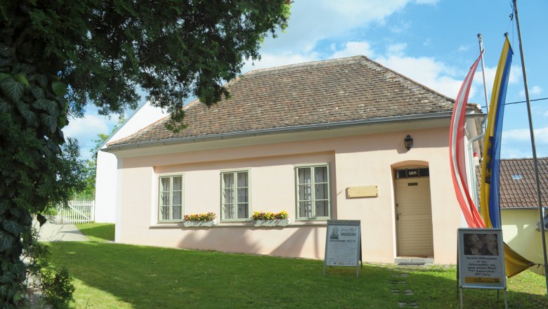 Pleyel Museum, © Prof. A. Ehrentraud A small, pink-painted building with a shingle roof, surrounded by trees and flags, with signs in front of the entrance.