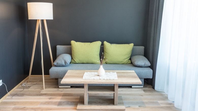 Living room example in the wine lodge, © KASTNER NEW MEDIA - kastner.tv Modern living room with gray sofa, green cushions, wooden lamp and coffee table in front of a dark wall.
