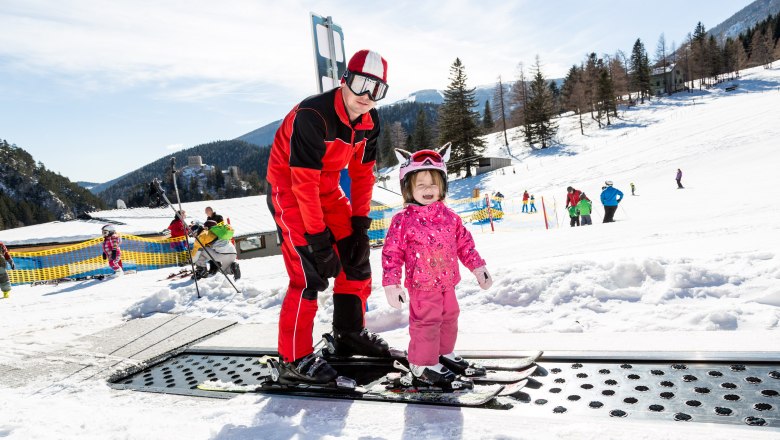 Learn to ski at the foot of the Schneeberg, © NÖ Schneebergbahn, Franz Zwickl A ski instructor helps a small child to ski on a conveyor belt in the snow.