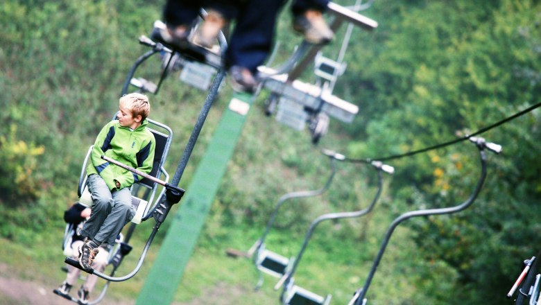 Lift ride up the Muckenkogel, © weinfranz.at A boy in a green jacket sits on a chair lift, surrounded by green nature.
