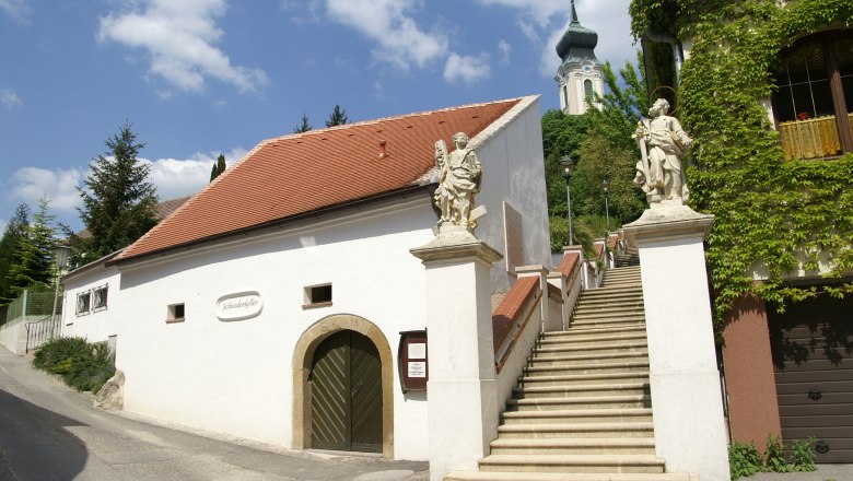 Swedish cellar, © Stadtgemeinde Mistelbach/Mag. Mark Schönmann White building with red roof and staircase with statues, church in the background.