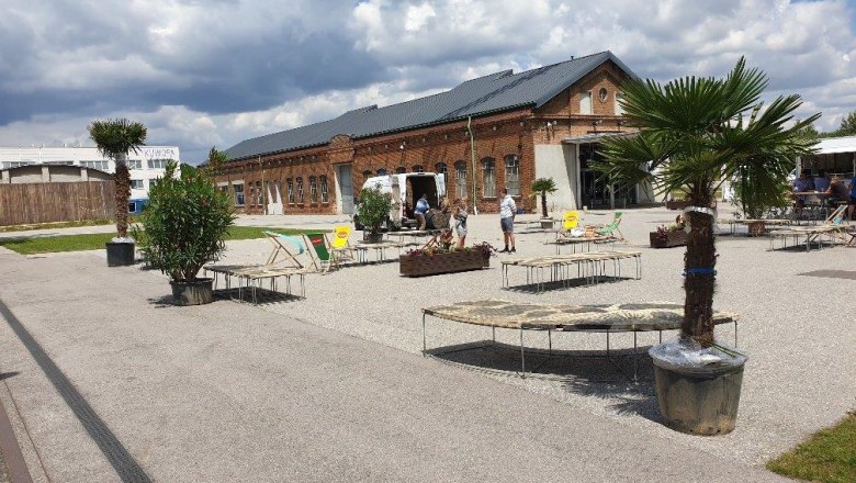Korneuburg-donau-werftbad-gastro-chill-area-palmen-c-sefko, © SEFKO Outdoor area with palm trees, sun loungers and a brick building in the background.
