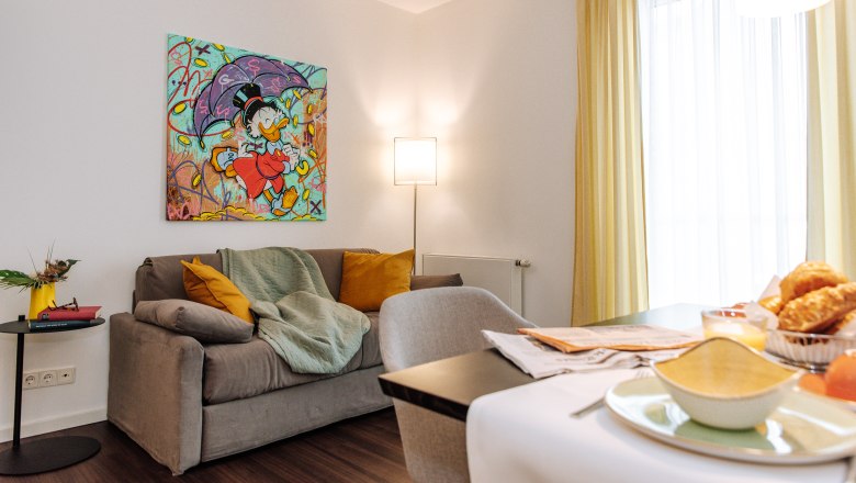Apartment HEINhotel vienna airport, © HEINhotel vienna airport Living room with gray sofa, colorful comic art on the wall, yellow curtains and breakfast table.