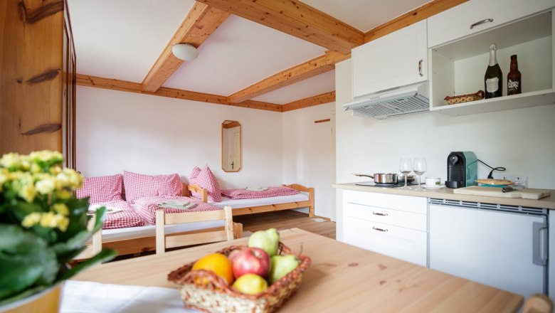 Cozy vacation apartment with wooden furniture, a small kitchen and a bed with red and white checkered pillows. There is a fruit basket on the table.