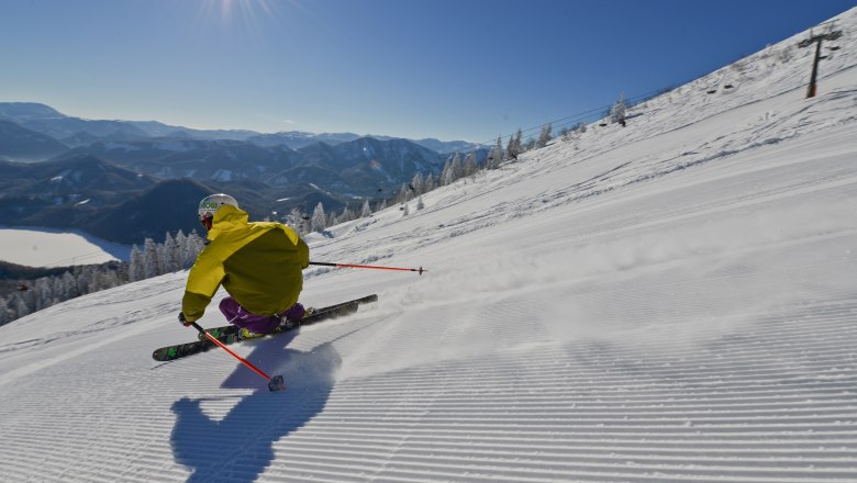 Skiers on a piste with a view of the Erlaufsee and mountains in the background.