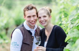 A smiling couple in a vineyard, the man wearing a vest, the woman holding a wine glass.