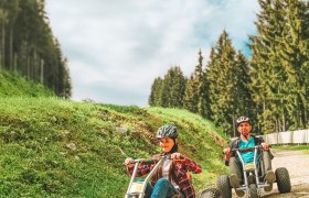 Two people ride mountain carts on a gravel road, surrounded by trees and blue sky.