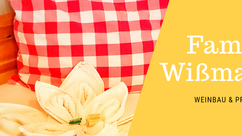 Red cushion with checked pattern and flower-shaped towel on a bed. Text: Wißmann family, winegrowing & private rooms.