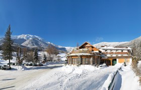 Winter landscape with Hotel Forellenhof in front of snow-covered mountains and a blue sky.
