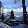 Snow-covered terrace with wooden benches and tables, surrounded by trees and mountains in the background.