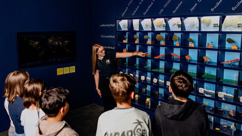 A group of young people watch a woman pointing at a wall of diagrams and models during a guided tour.