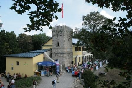 A yellow building with a stone tower, surrounded by trees and people gathering outside.