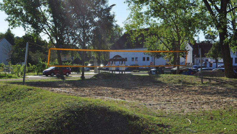 An empty volleyball court with a net, surrounded by trees and parked cars in the background.