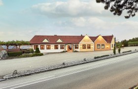 Exterior view of a winery with a yellow building and red roof, surrounded by vines and a road in the foreground.