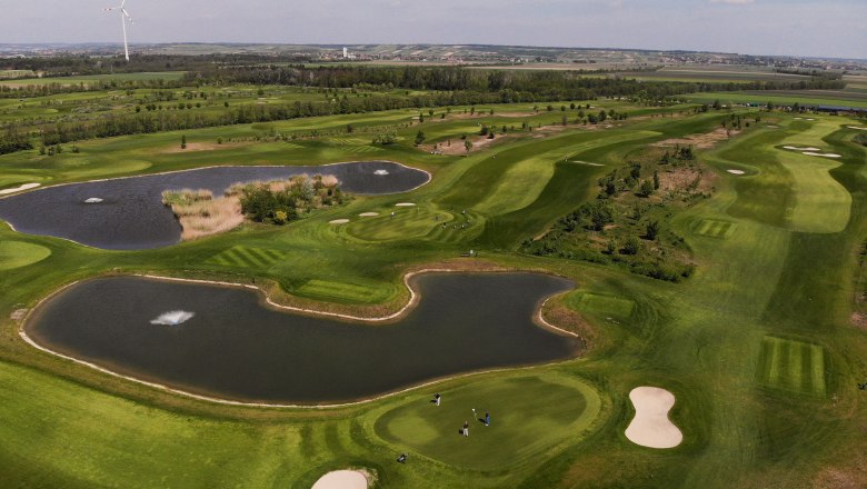 Aerial view of a golf course with ponds and wind turbine in the background.