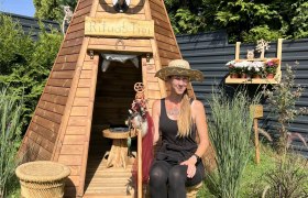 Woman in a straw hat sits in front of a wooden tepee in the garden.