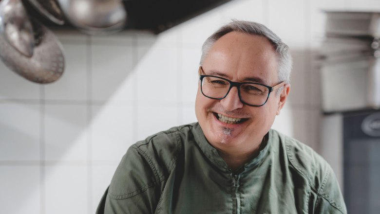 A smiling chef in a green jacket stirs a pot in a professional kitchen.