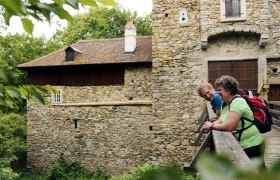 Two people lean against a railing in front of an old stone wall.