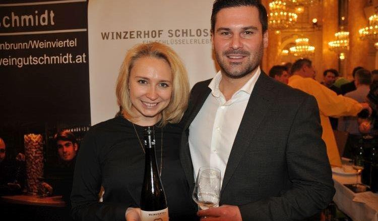 A couple holds a wine bottle and a wine glass in front of a banner with the inscription 'Winzerhof Schlosser'.