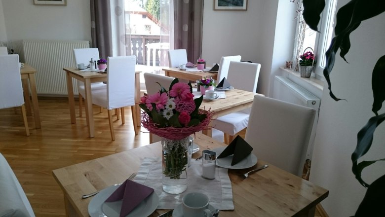 A bright breakfast room with wooden tables, white chairs and flower arrangements on the tables.