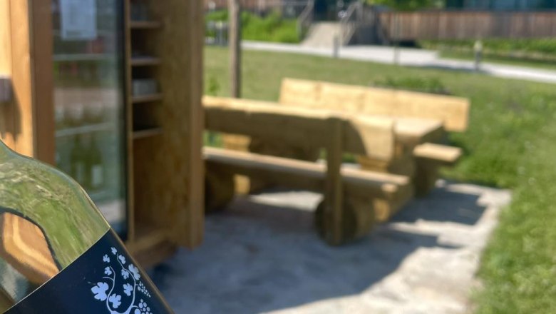 A bottle labeled 'Spritzer rocks' in front of a rest area with wooden benches and an outdoor fridge.