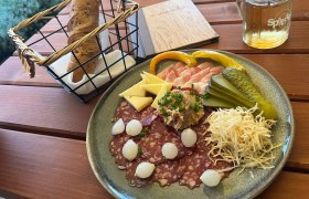 A plate with cold cuts, cheese, gherkins and onions, next to it a basket of bread and a drink on a wooden table.