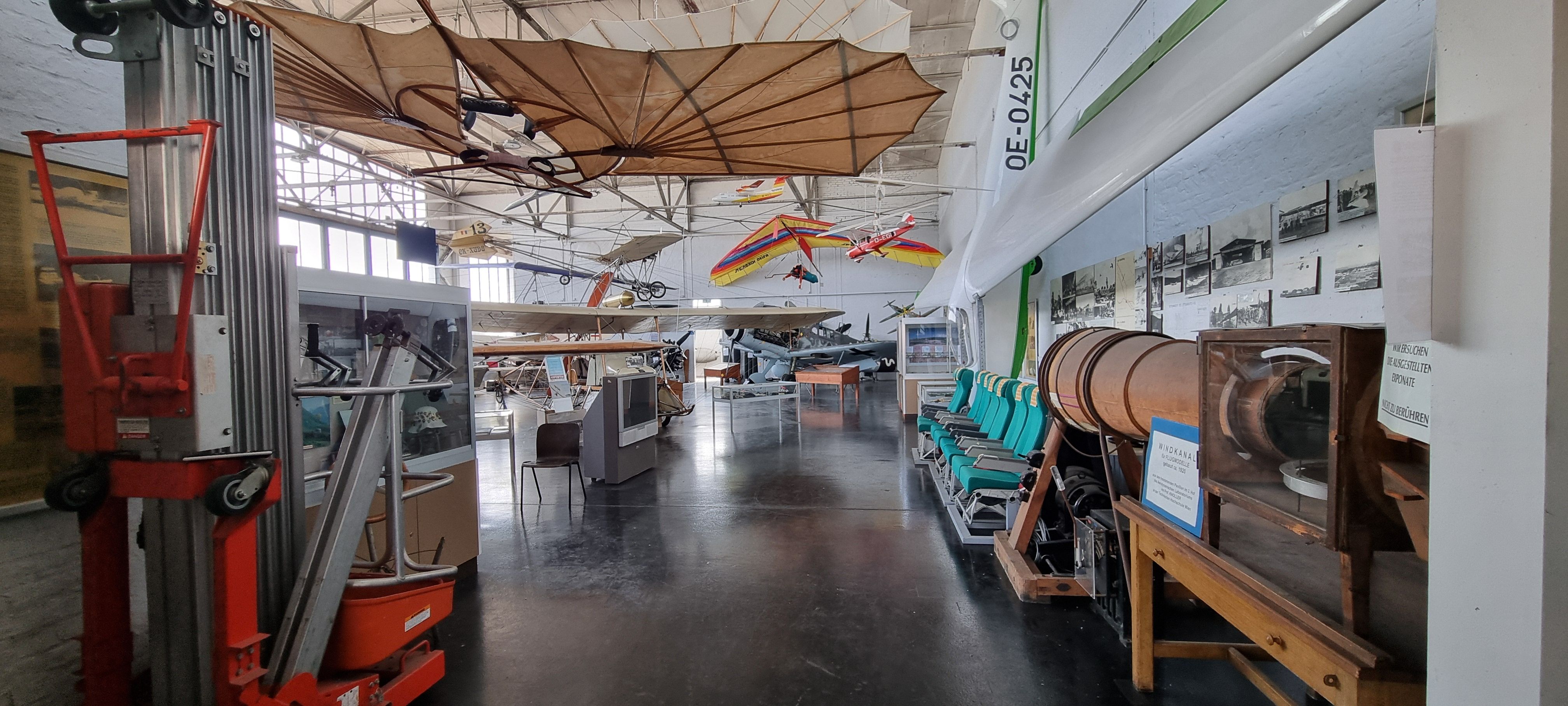 Large hall with exhibits hanging from the ceiling, airplane seats, information displays and televisions.