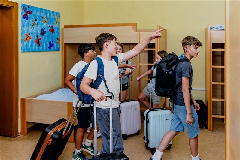 Group of children with suitcases in a dormitory with bunk beds.