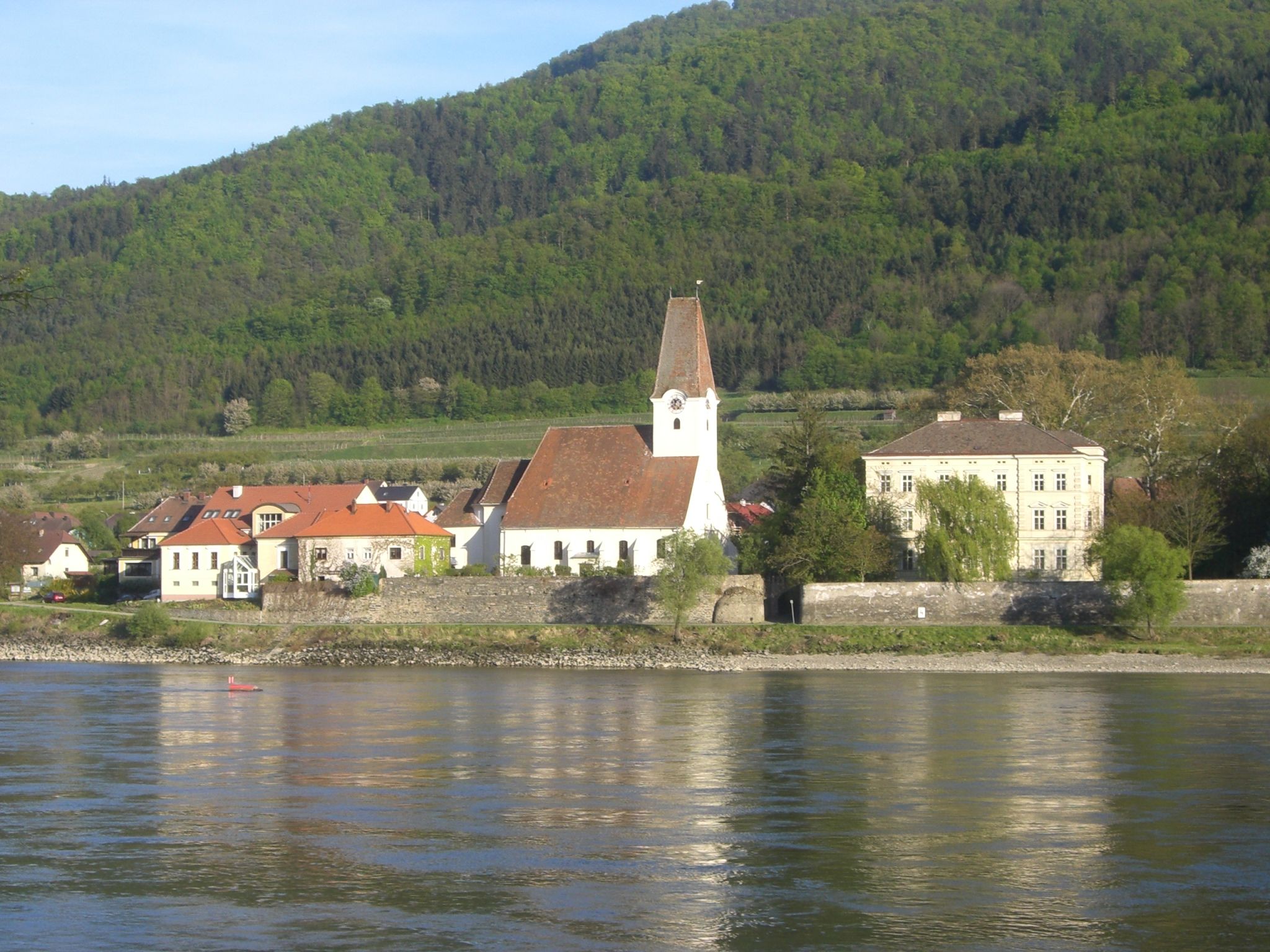 Hofarnsdorf parish church with surrounding buildings and wooded hill in the background, taken from the Danube.