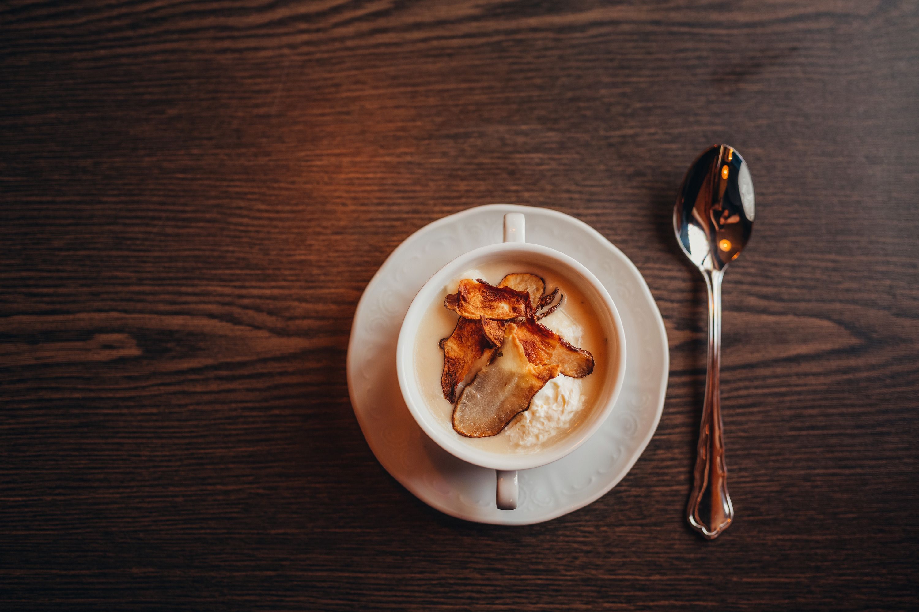 A cup of Jerusalem artichoke soup with chips on a wooden table, a spoon next to it.