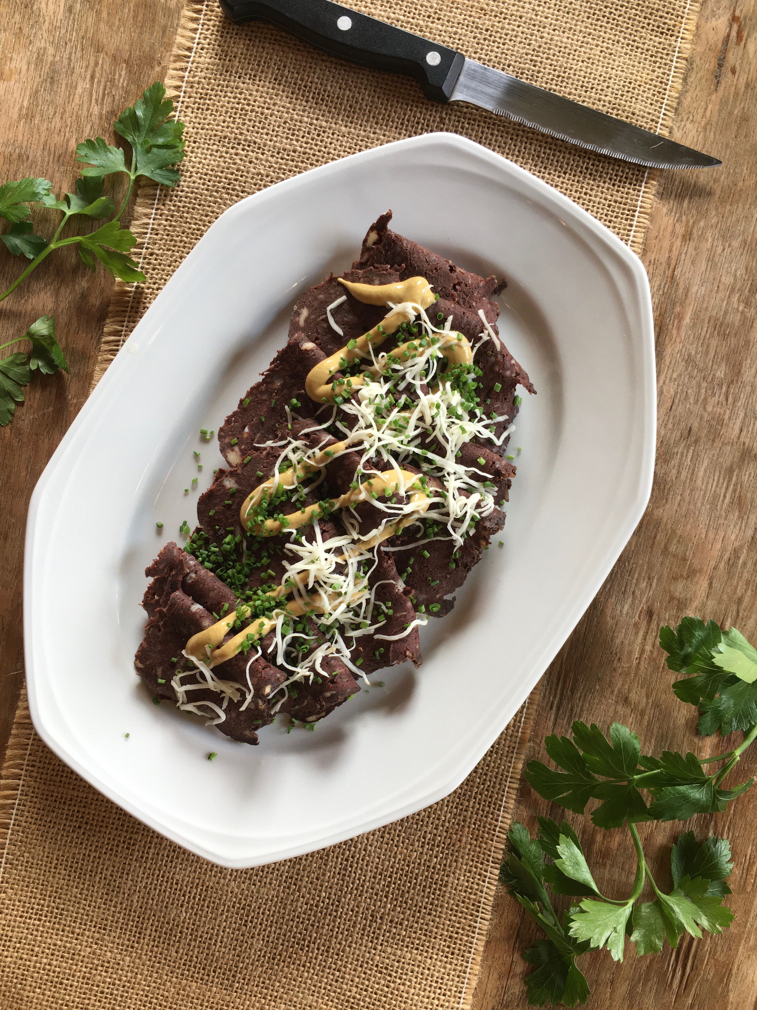 A plate of Blunzenbrot, garnished with mustard, chives and grated cheese, on a wooden table with parsley and a knife next to it.
