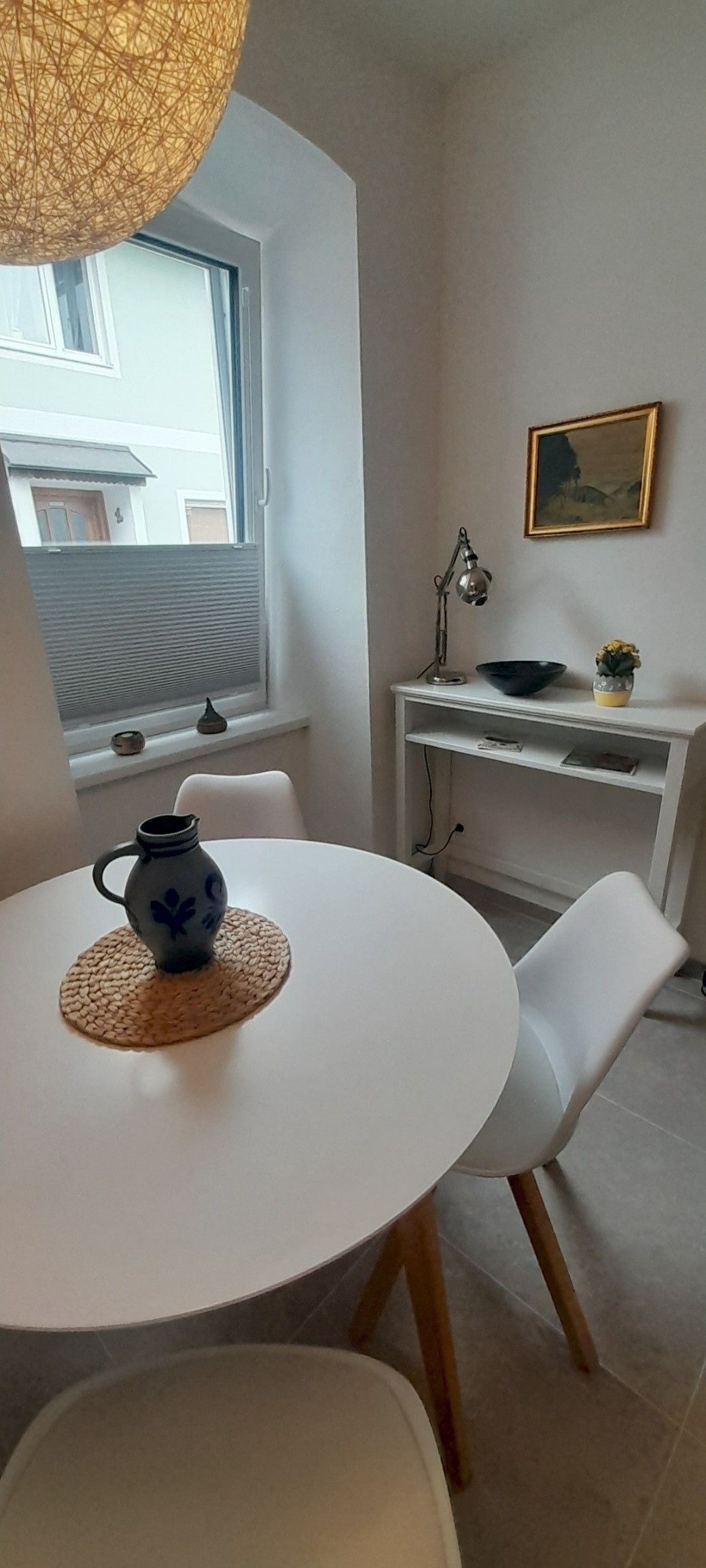 A small dining area with a round white table, two white chairs and a decorative vase. In the background, a window and a small table with a lamp.
