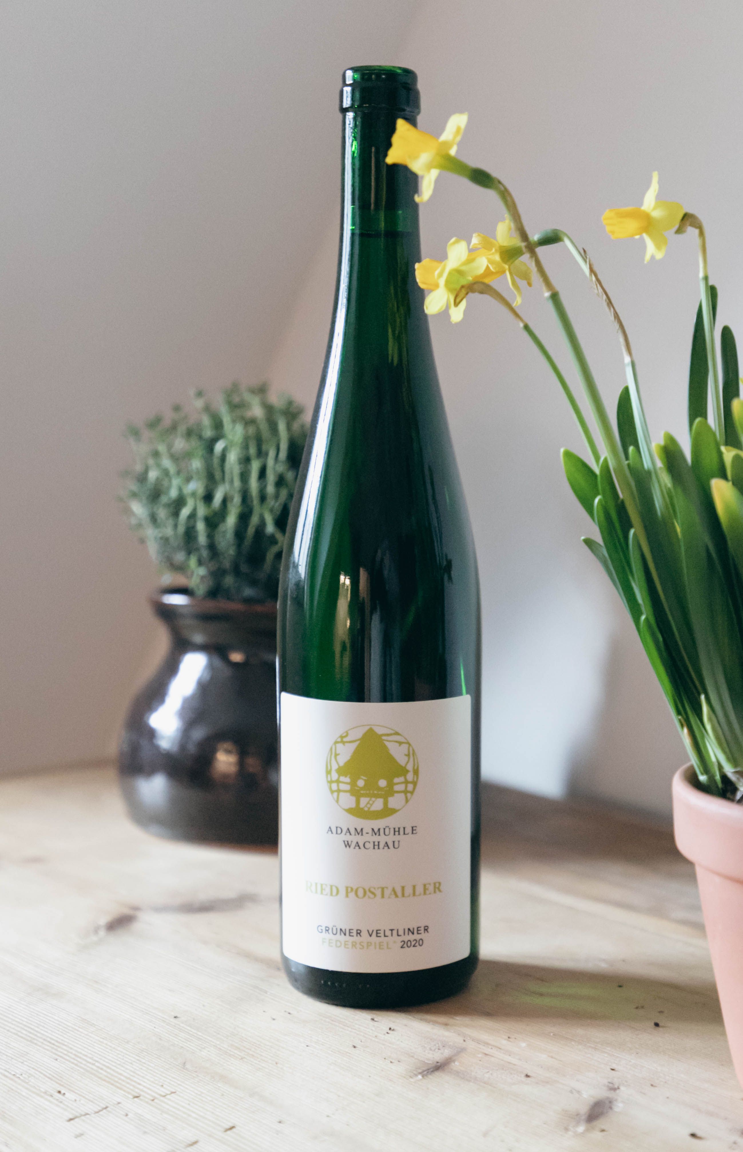 A bottle of Grüne Veltliner wine sits on a table next to yellow flowers and a pot of herbs.