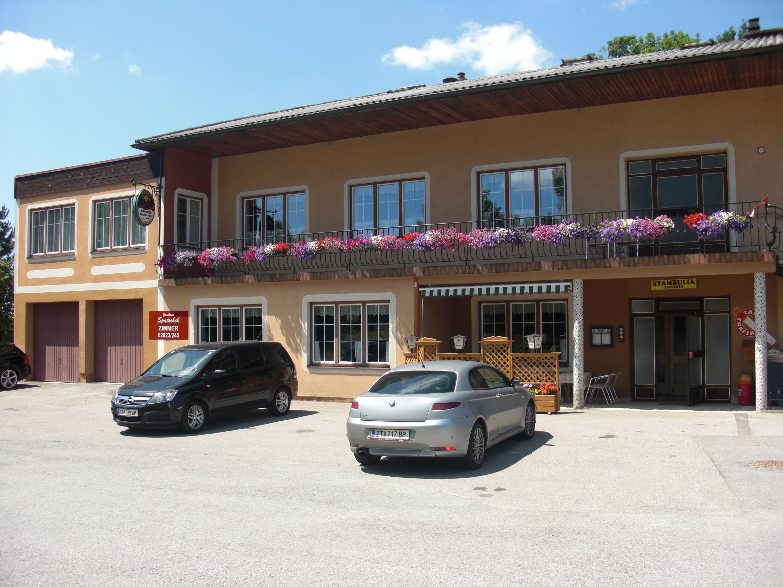 Inn with balcony and flowers, two parked cars in front.