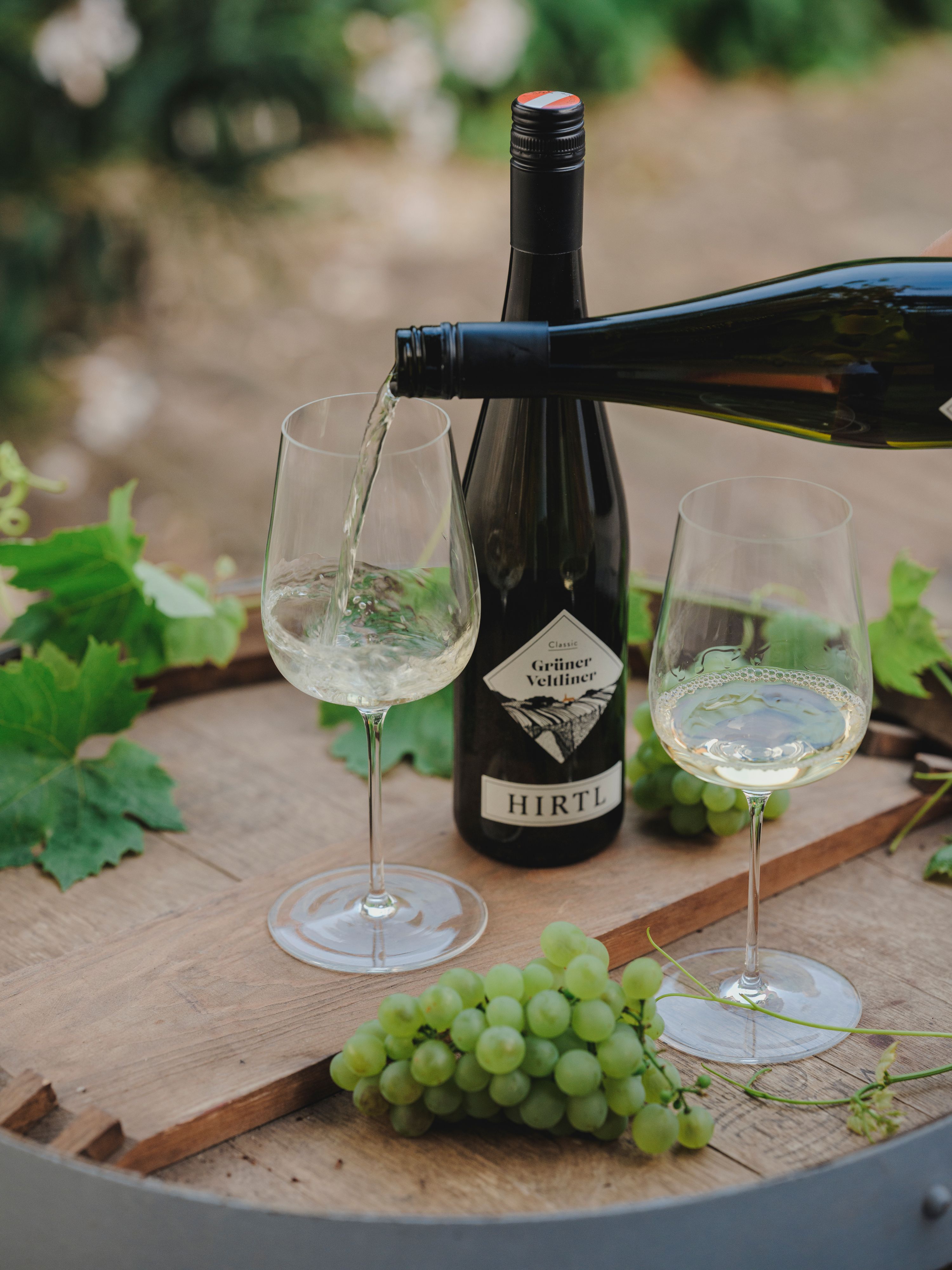 White wine is poured into a glass, next to it a bottle of Grüne Veltliner from Hirtl and green grapes on a wooden table.