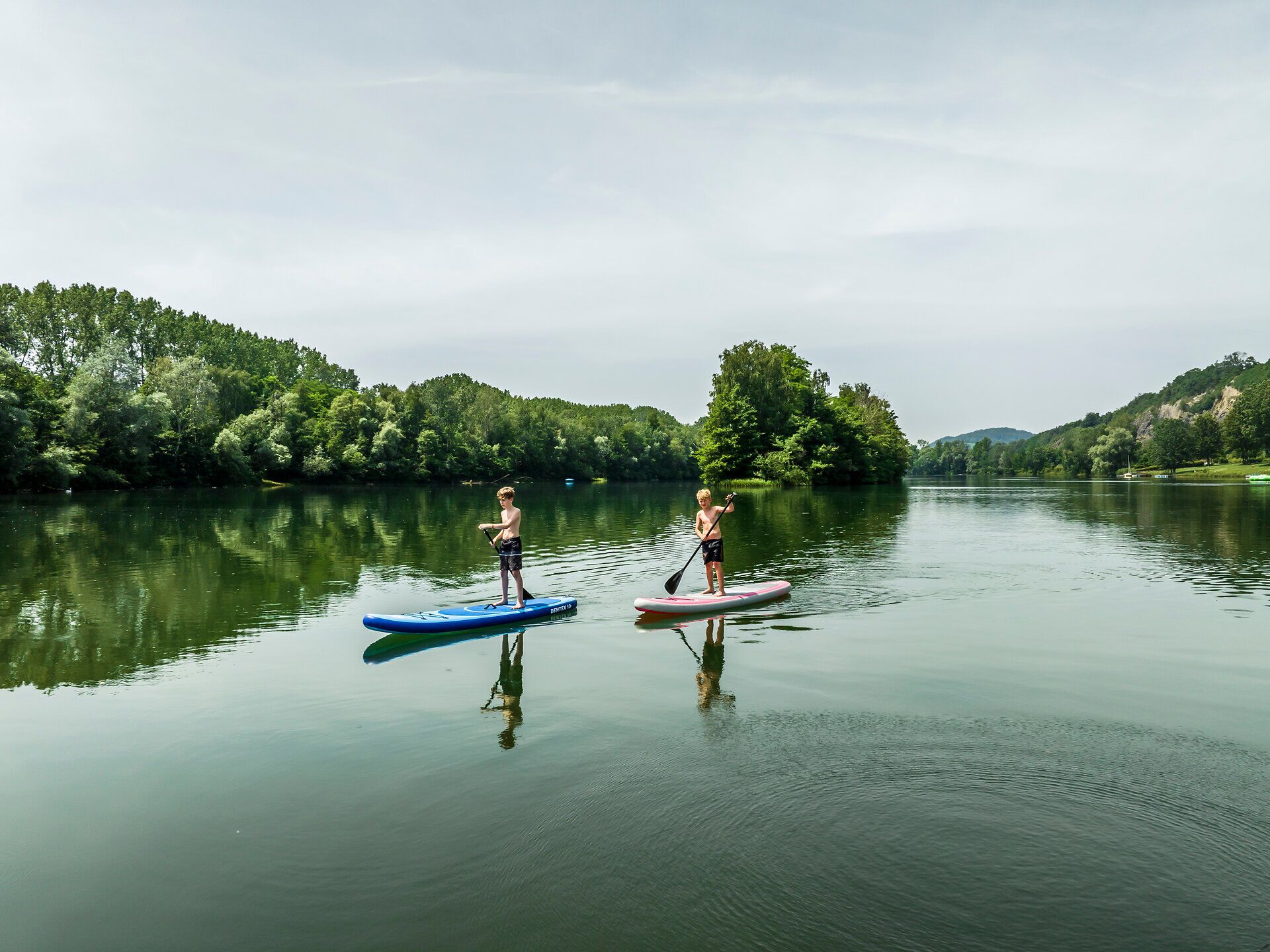 Three people on a pedal boat by the lake, bicycles on the jetty, green trees in the background.