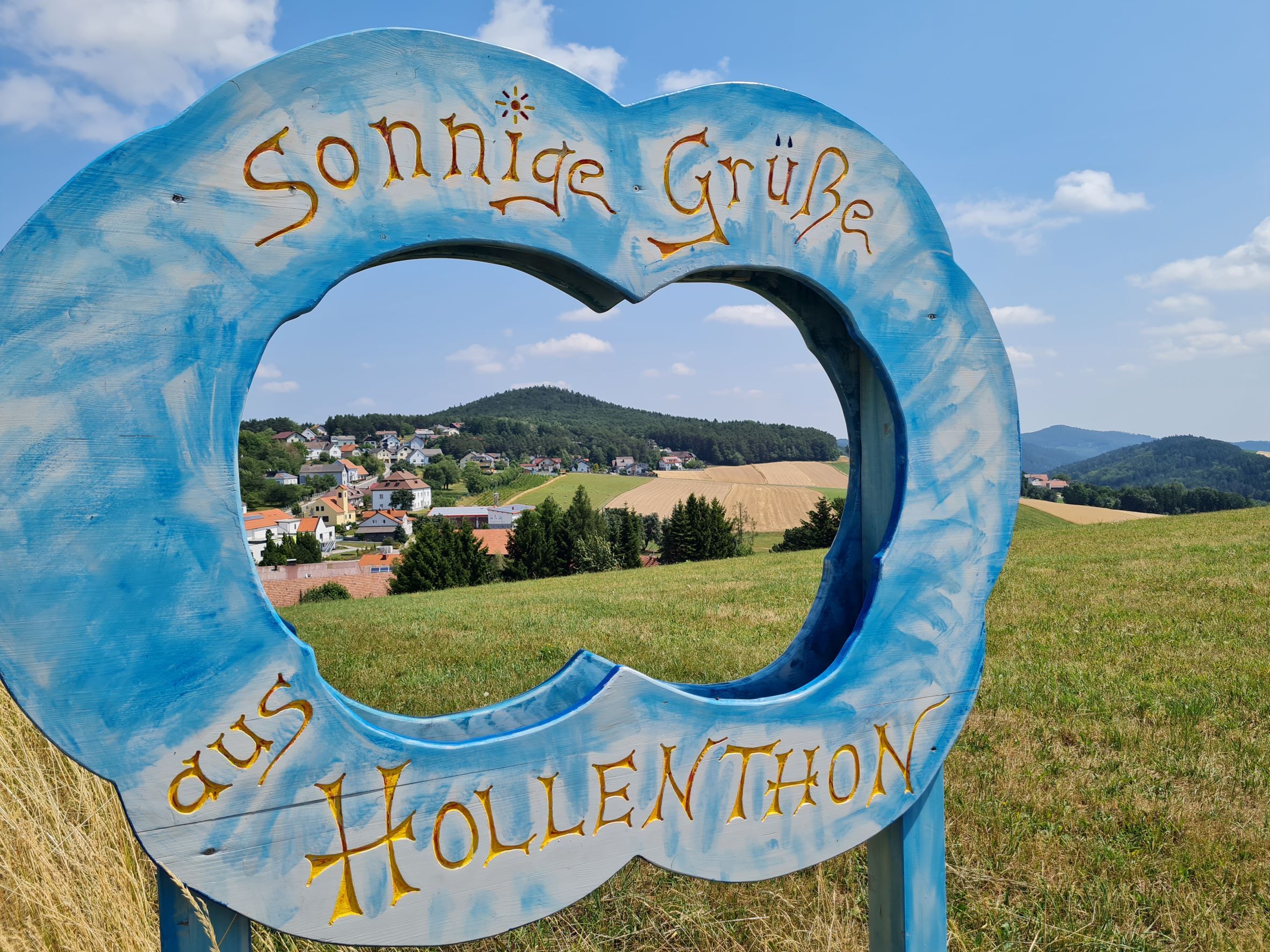 Blue wooden frame in the shape of a cloud with the lettering 'Sunny greetings from Hollenthon' in front of a landscape.