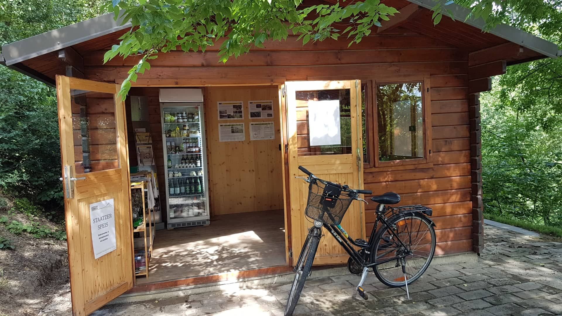 Small wooden house with open entrance, fridge with drinks and a bicycle in front.