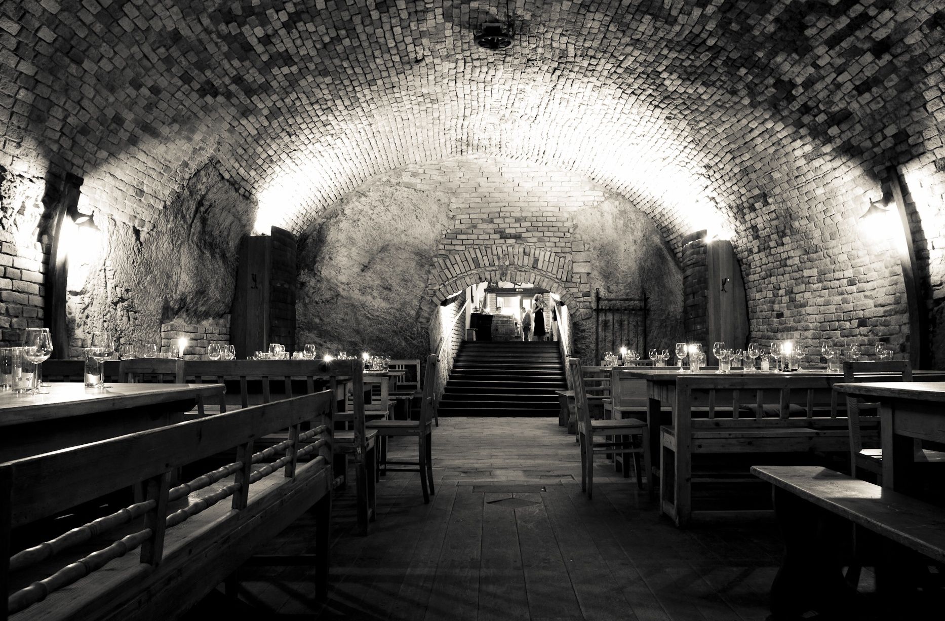 A rustic wine cellar with wooden tables and benches, illuminated by wall lamps.