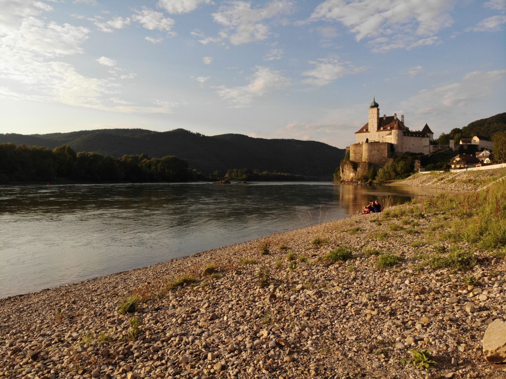 Pebble beach on the Danube with castle in the background at sunset.