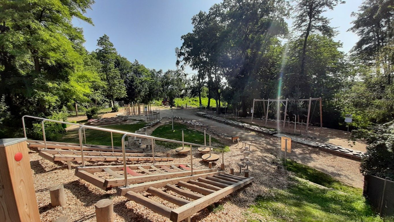 A motor skills park with various wooden elements and climbing equipment, surrounded by trees and green spaces.