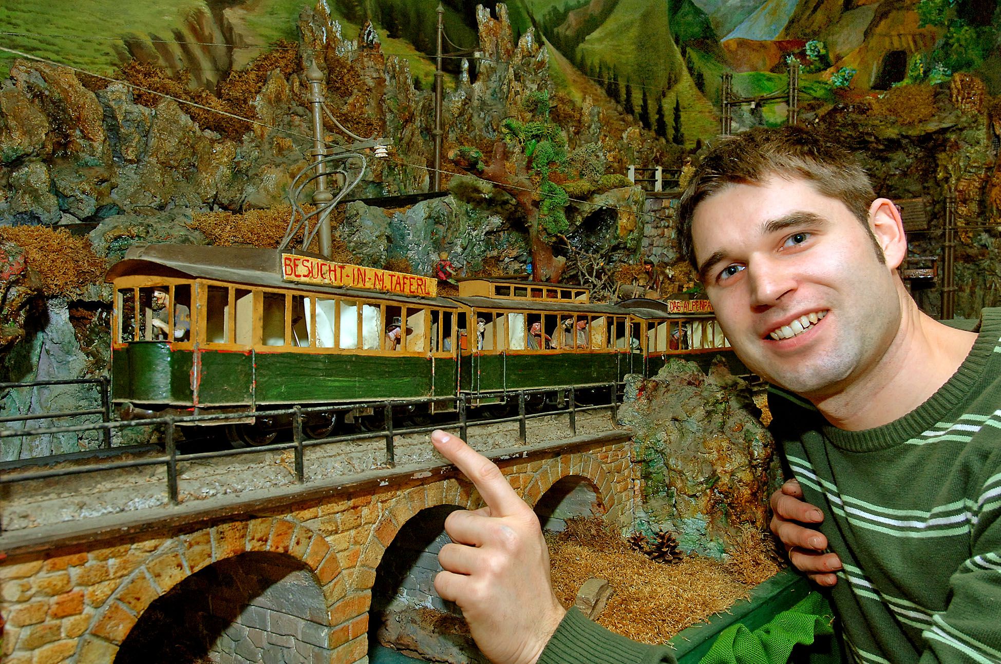 A man points to a model railroad in an alpine landscape.