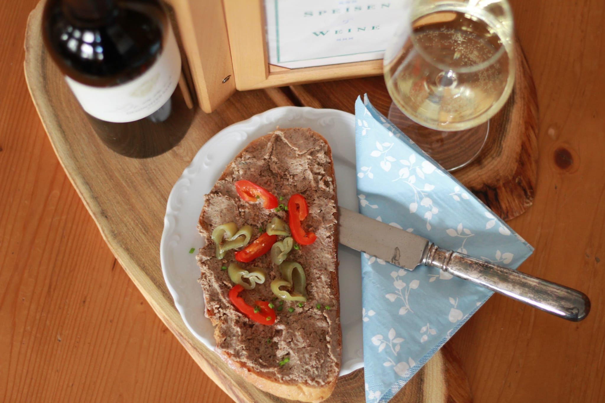 A plate with spread, peppers and a knife, next to it a glass of white wine and a bottle of wine on a wooden table.