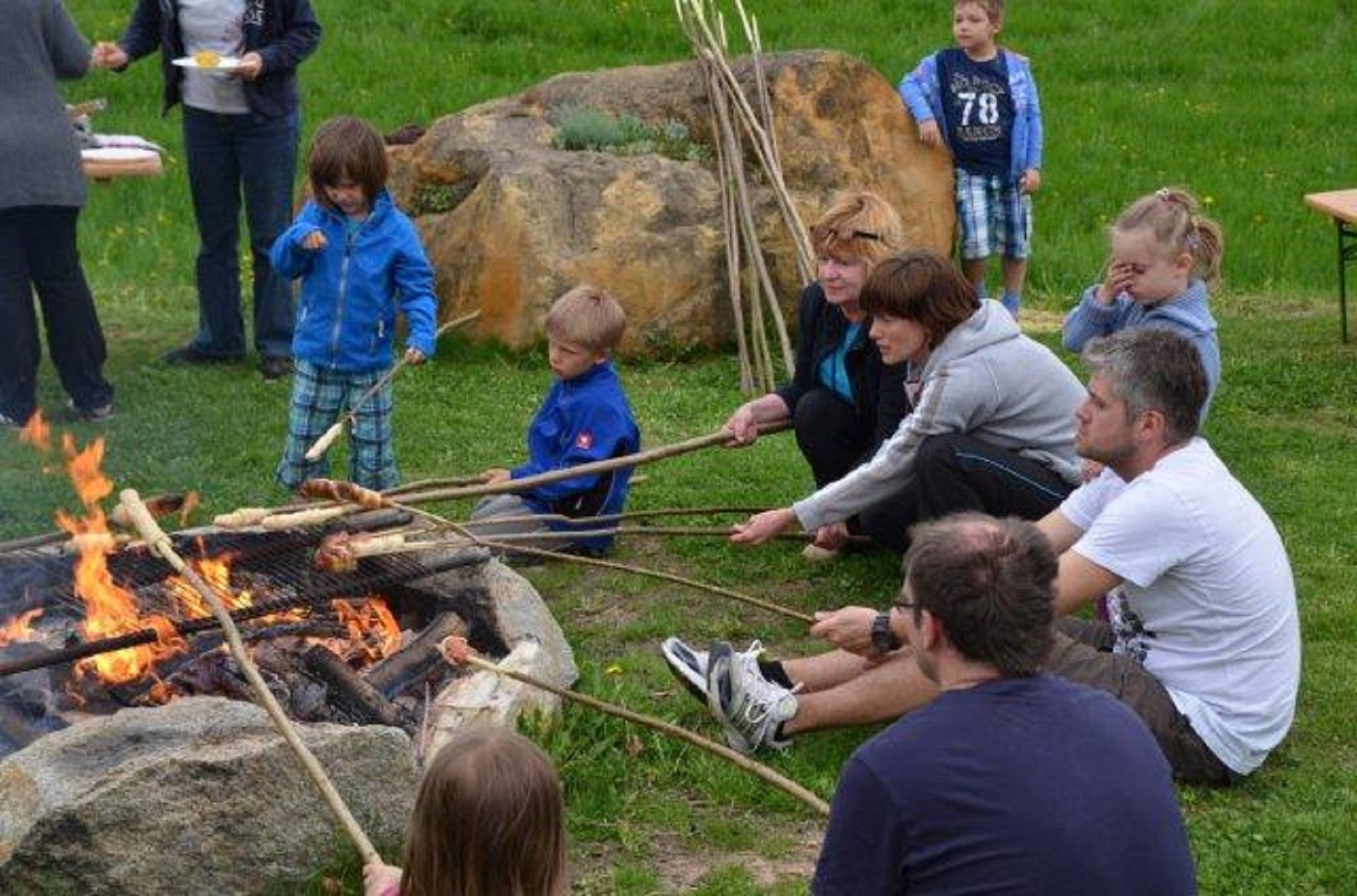 People sit around a campfire and roast bread on long sticks.