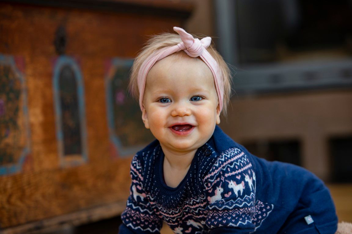 A smiling baby with a pink headband and blue sweater with a reindeer pattern.