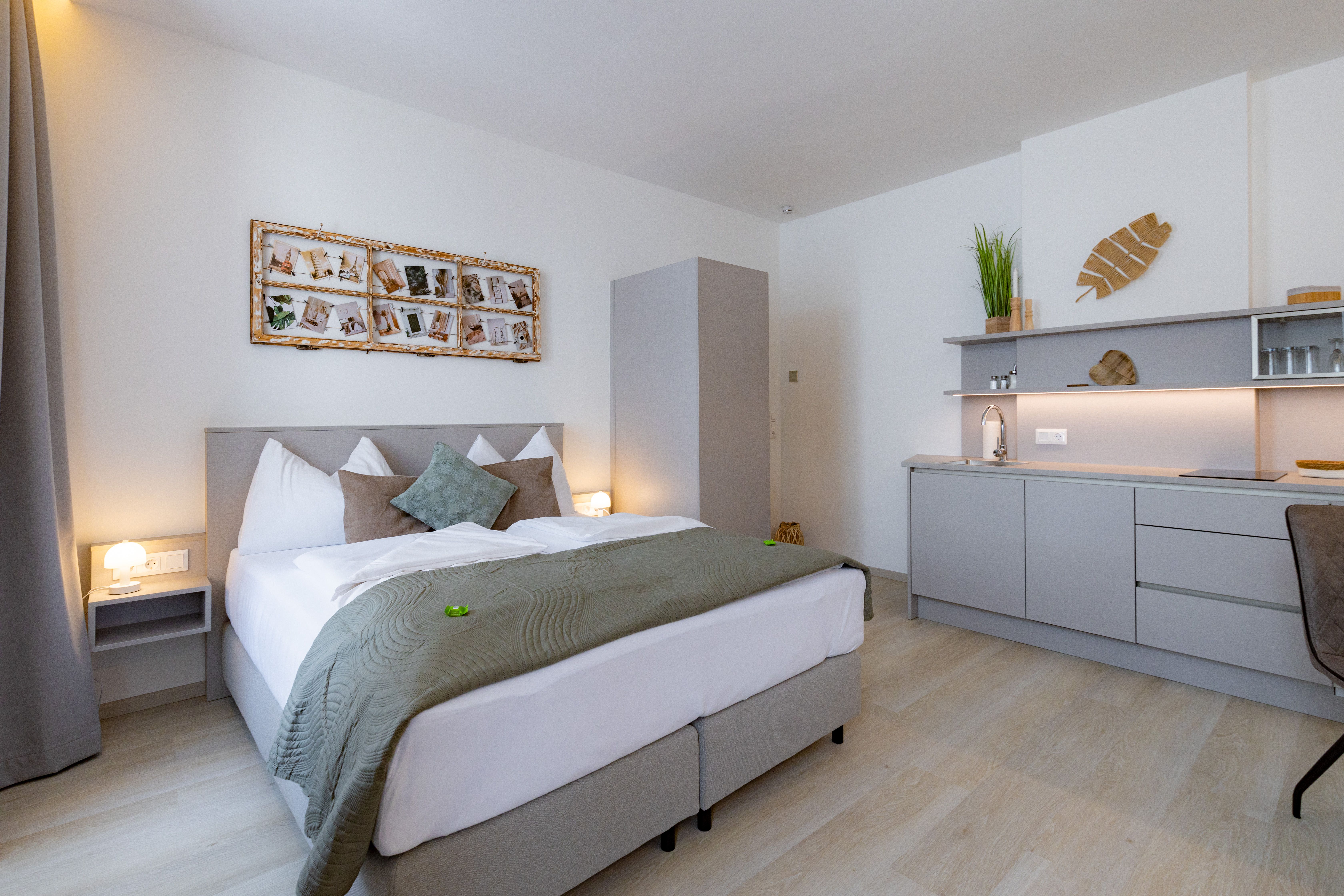 Modern bedroom with double bed, gray bed linen, wall decoration and small kitchenette.