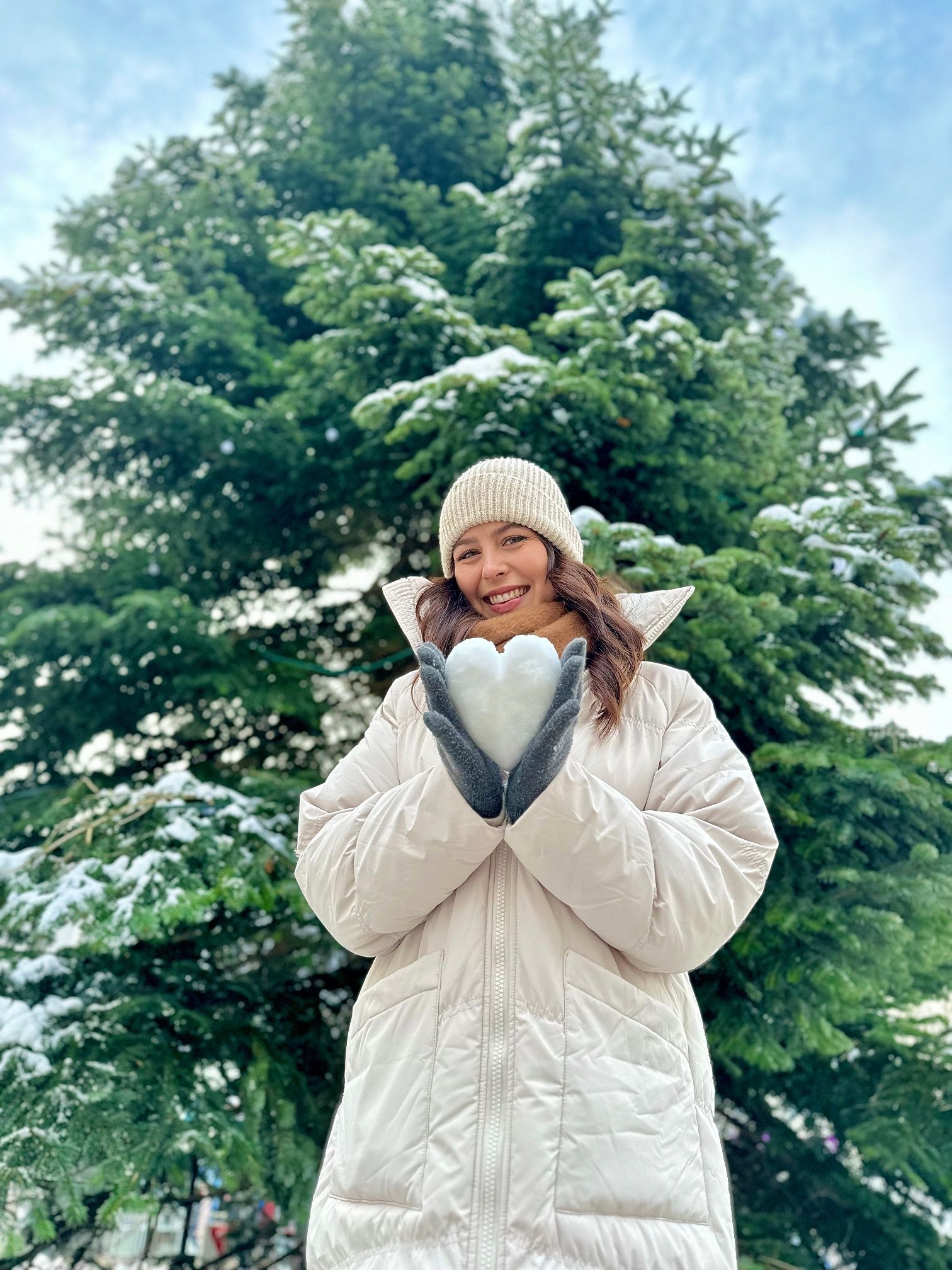 Woman in a white coat holds a heart made of snow in front of a snow-covered tree.