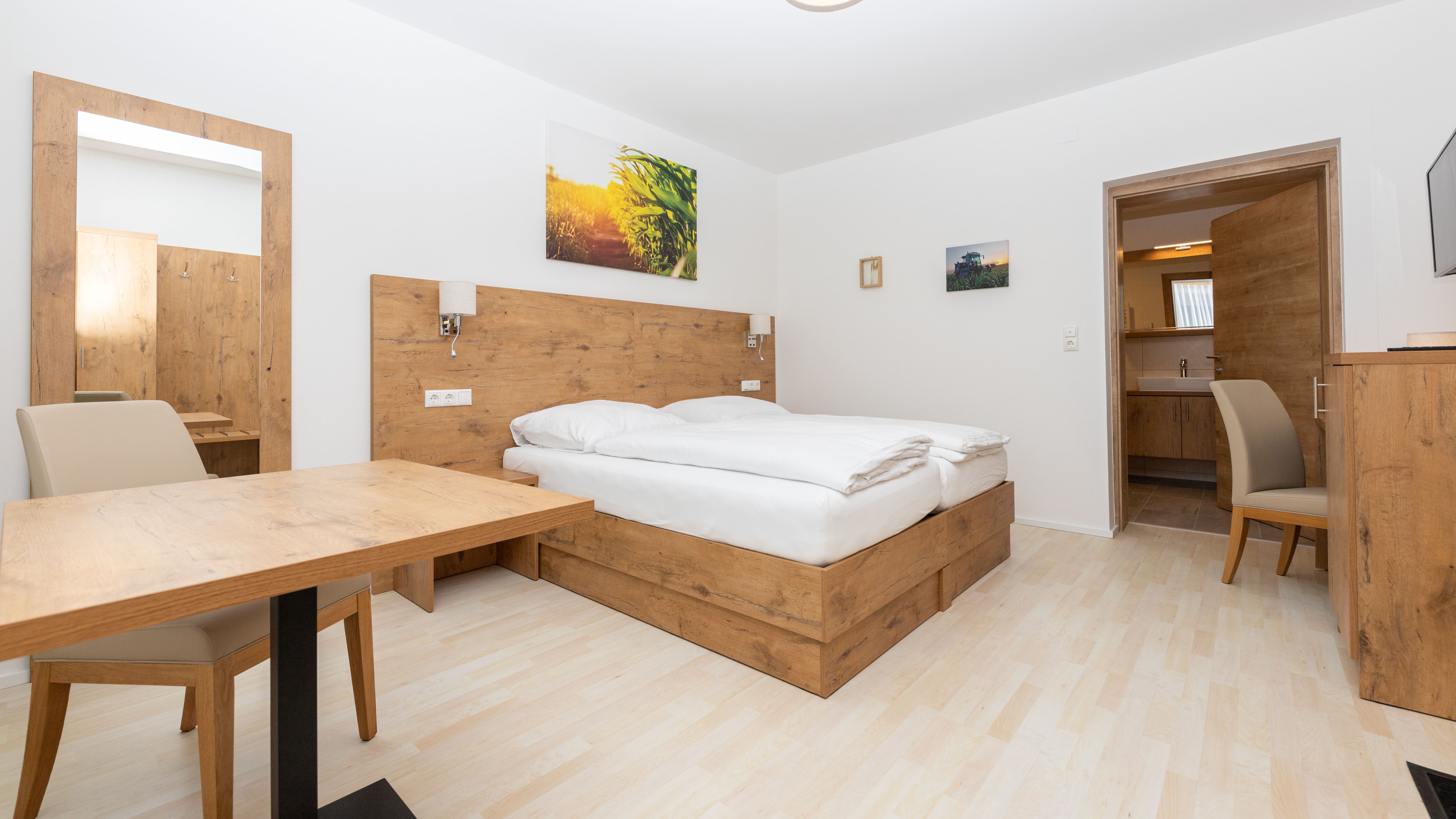 Modern bedroom with wooden furniture, double bed, table and chair. A mirror and pictures on the wall, door leading to a bathroom.