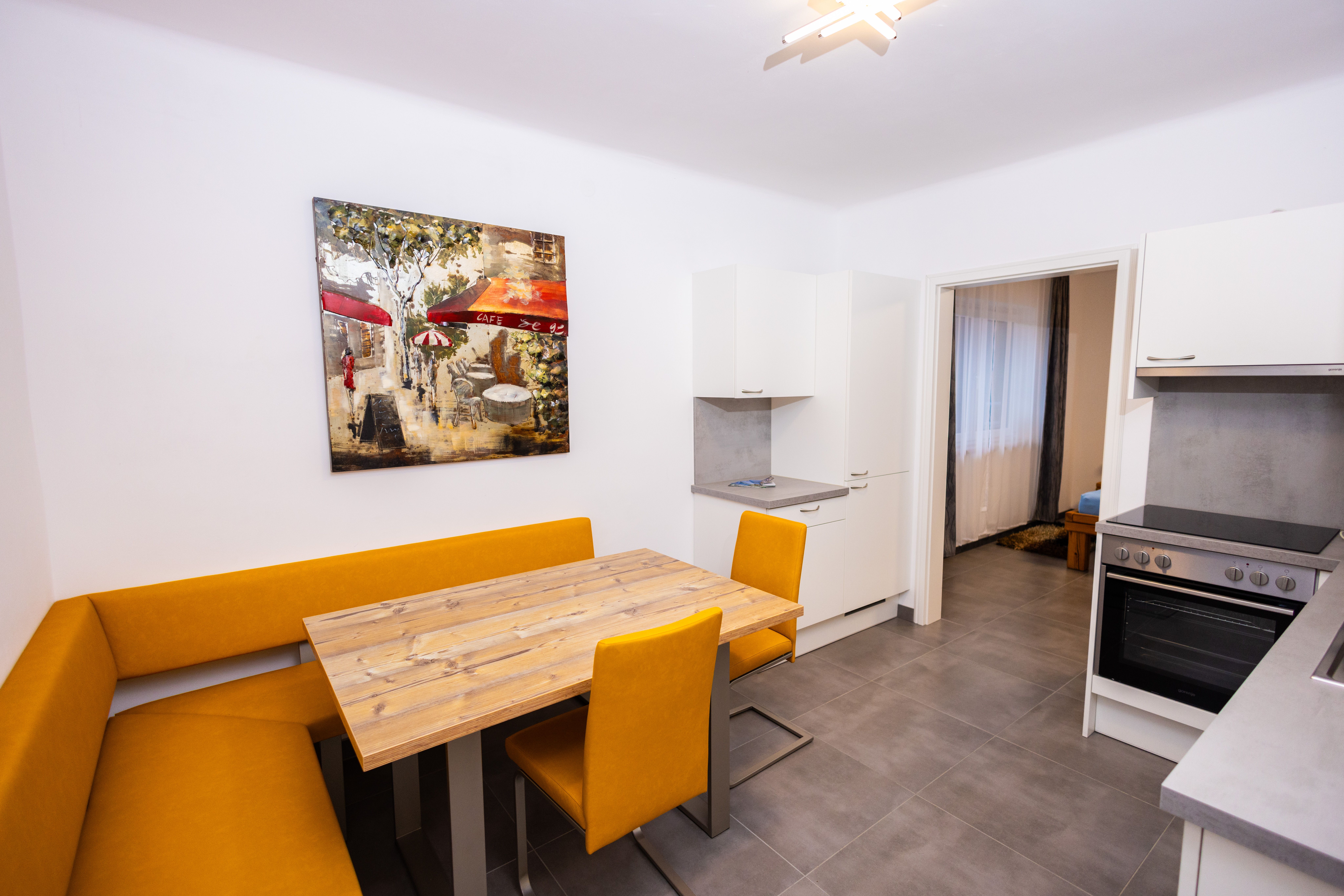 Modern kitchen with yellow corner bench, wooden table, white cupboard and stove. A painting of a café hangs on the wall.